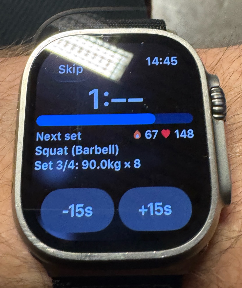 Photo of an Apple Watch Ultra on a wrist showing the Hevy rest timer dimmed, with the time shown as 1:-- above a progress bar, the next set Squat (Barbell) 3 of 4 at 90 kg for 8, and -15s and +15s buttons