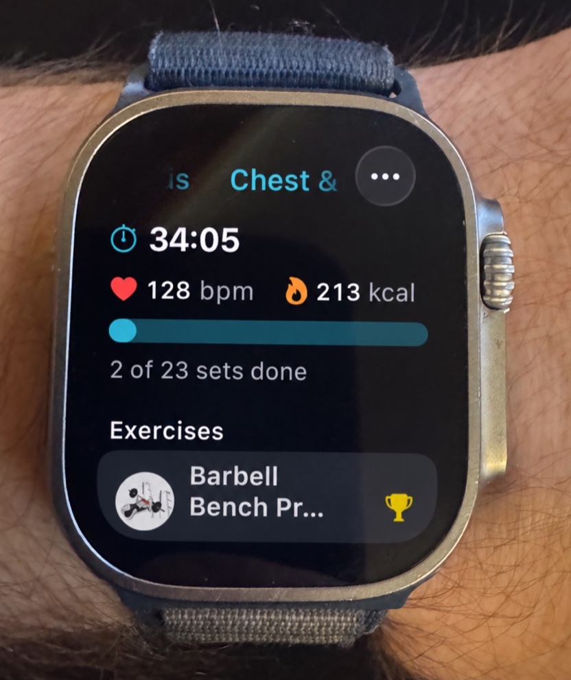 SetGrip workout screen on an Apple Watch Ultra on a wrist, showing a Chest and Triceps workout with 34:05 elapsed, 128 bpm heart rate, 213 kcal and 2 of 23 sets done